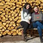 Two women sitting on a bench with a laptop, smiling outdoors against stacked logs.